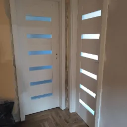 Dwa białe, przeszklone drzwi wewnętrzne z poziomymi szybami, zamontowane w surowym otworze, na tle brązowej podłogi ułożonej w jodełkę.