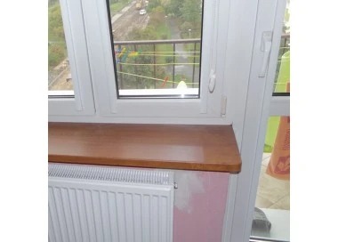 Wykończony parapet z drewna pod białym oknem PCV. Widok na zewnątrz z suszącym się praniem. Grzejnik pod parapetem. Fragment ściany z śladami po remoncie.