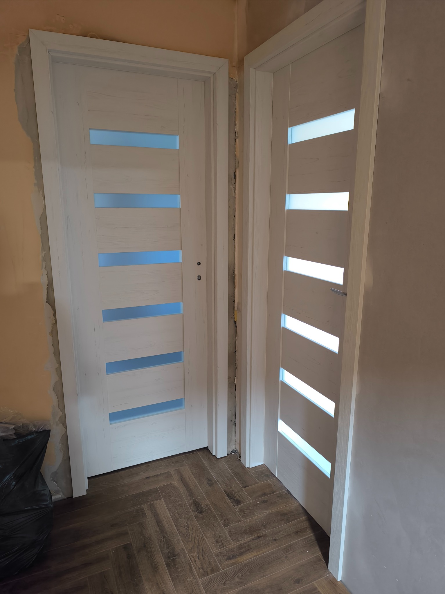 Dwa białe, przeszklone drzwi wewnętrzne z poziomymi szybami, zamontowane w surowym otworze, na tle brązowej podłogi ułożonej w jodełkę.