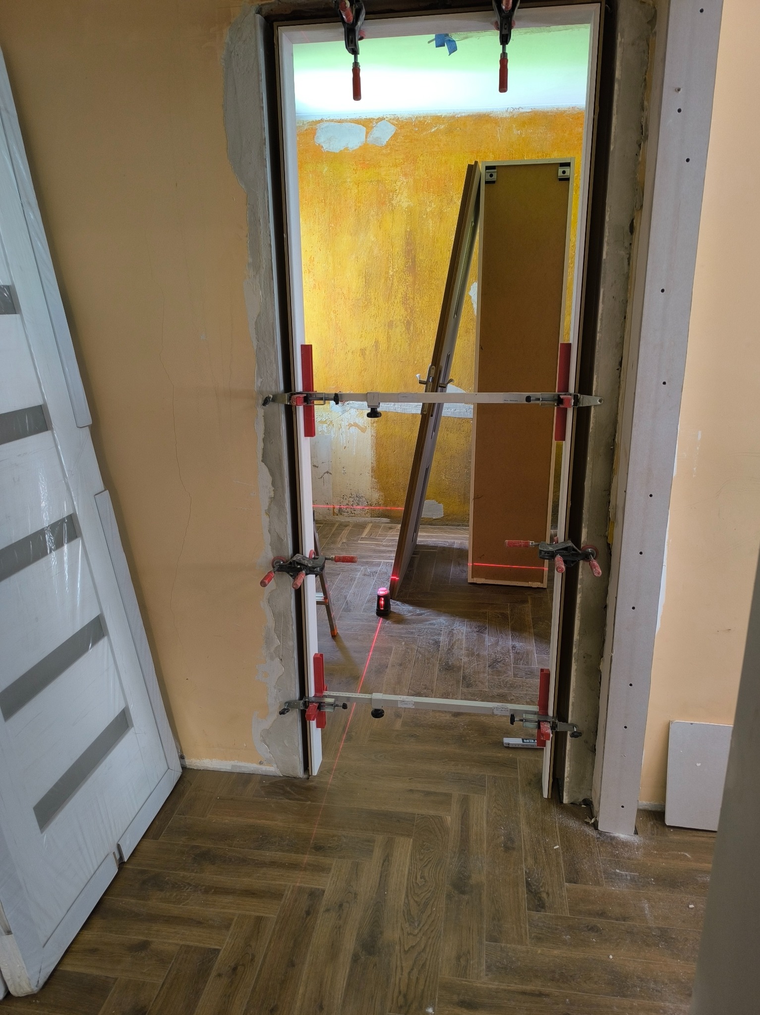 Montaż ościeżnicy z użyciem poziomicy laserowej, widoczne klamry mocujące, fragment podłogi ułożonej w jodełkę, ściana w kolorze ciepłego beżu, w tle drzwi oparta o ścianę.