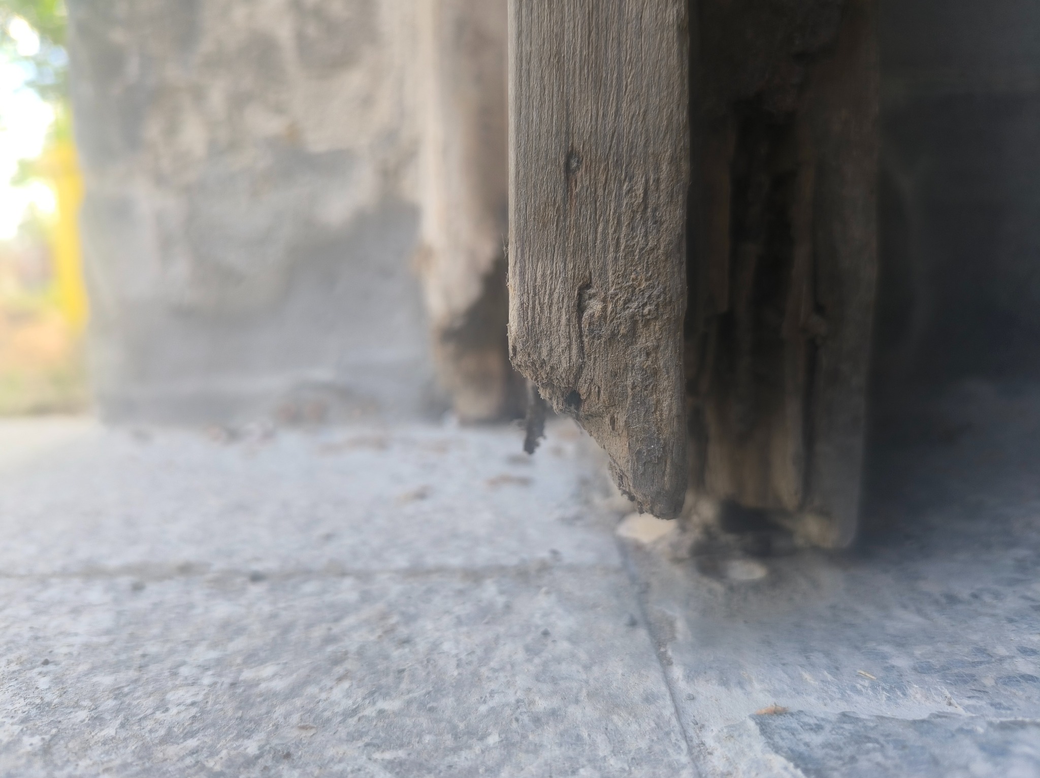 Niski kąt ujęcia fragmentu drewnianej ramy okiennej z widocznymi śladami zużycia i uszkodzeń, na tle szarego muru. Widoczne detale tekstury drewna i betonu.