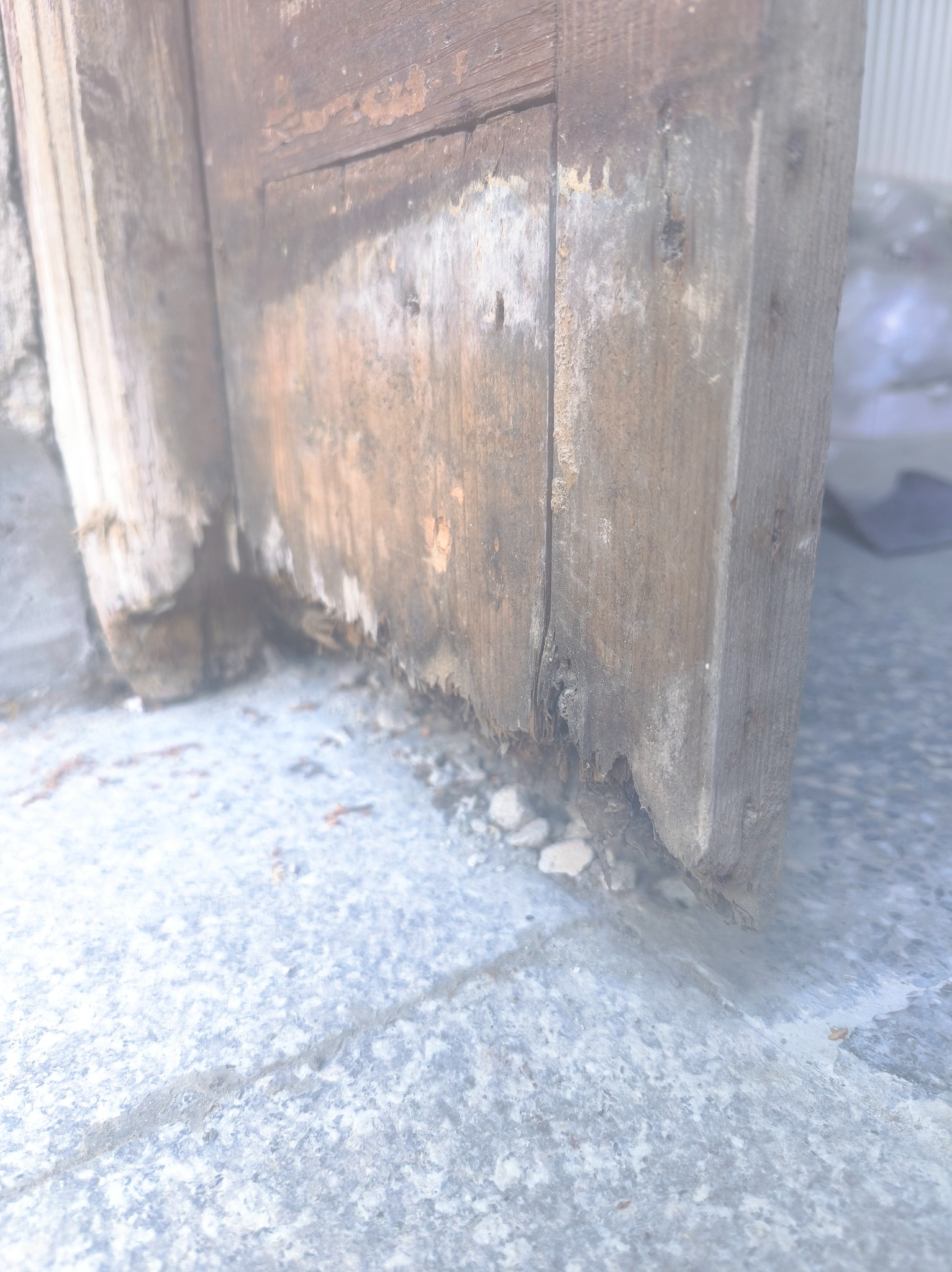 Uszkodzony, dolny fragment drewnianej ramy drzwiowej z widocznymi śladami zużycia i ubytkami, osadzony na szarej, kamiennej posadzce.