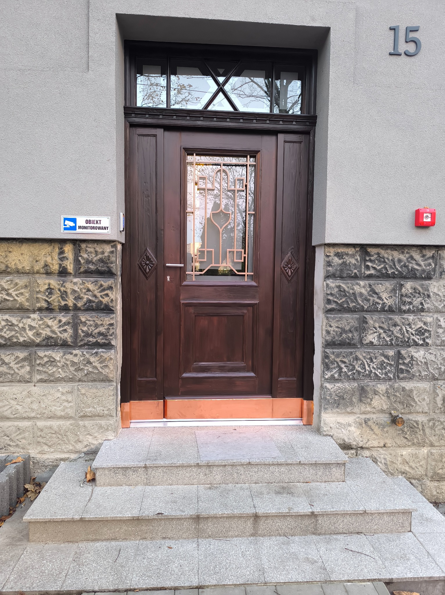 Ciemnobrązowe, odnowione drzwi wejściowe z szybą i ozdobną kratą, osadzone w kamiennej fasadzie budynku z numerem 15. Widoczne schody wejściowe i system monitoringu.