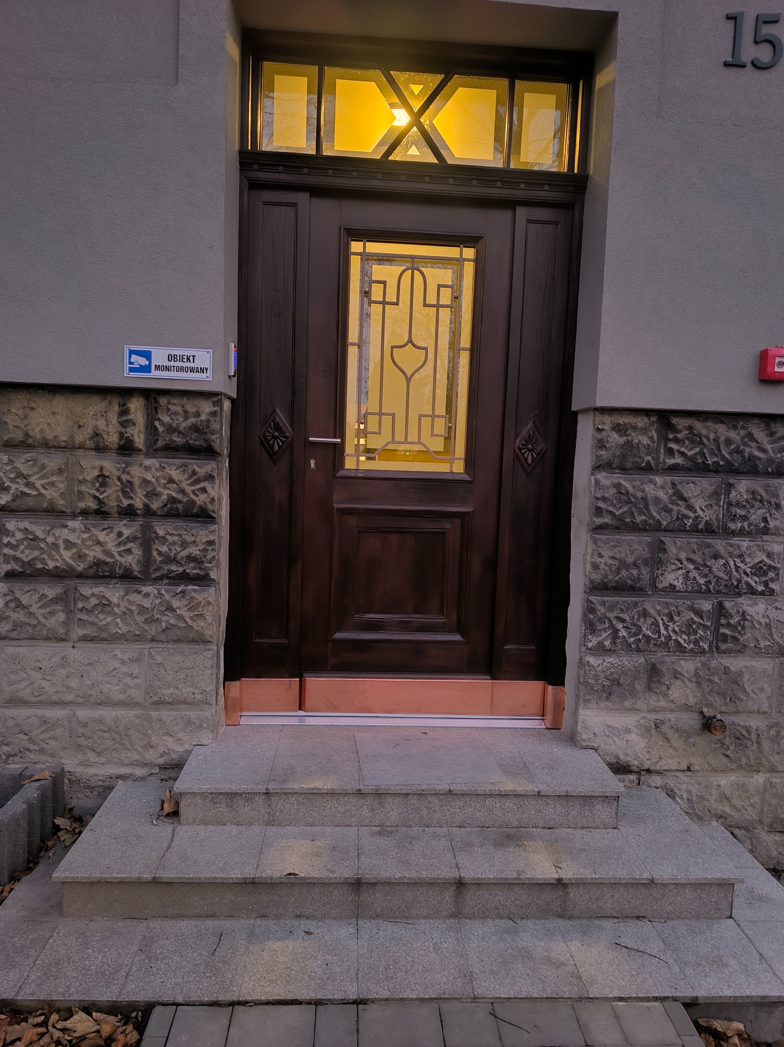 Ciemne, drewniane drzwi wejściowe z szybą i ozdobnymi elementami, oświetlone od wewnątrz, z numerem 15 na ścianie i schodami prowadzącymi do wejścia.