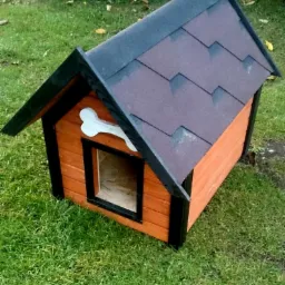 Drewniana buda dla psa z dachem krytym gontem bitumicznym, z dekoracyjną kością nad wejściem, wykonana w Kłobucku. Pomarańczowe ściany, czarne wykończenia.