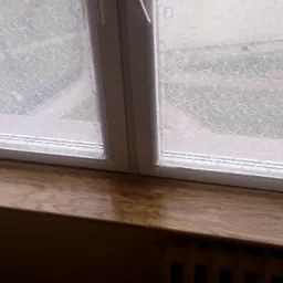 Jasny, drewniany parapet pod oknem z firanką w delikatny wzór. Widoczny fragment grzejnika. Ujęcie z bliska, skupione na detalu wykończenia.