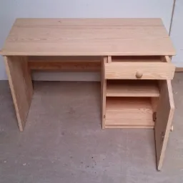 Jasny, drewniany biurko z wysuwaną szufladą i otwartą szafką po prawej stronie, ukazujące wnętrze z półkami. Widoczne naturalne usłojenie drewna.