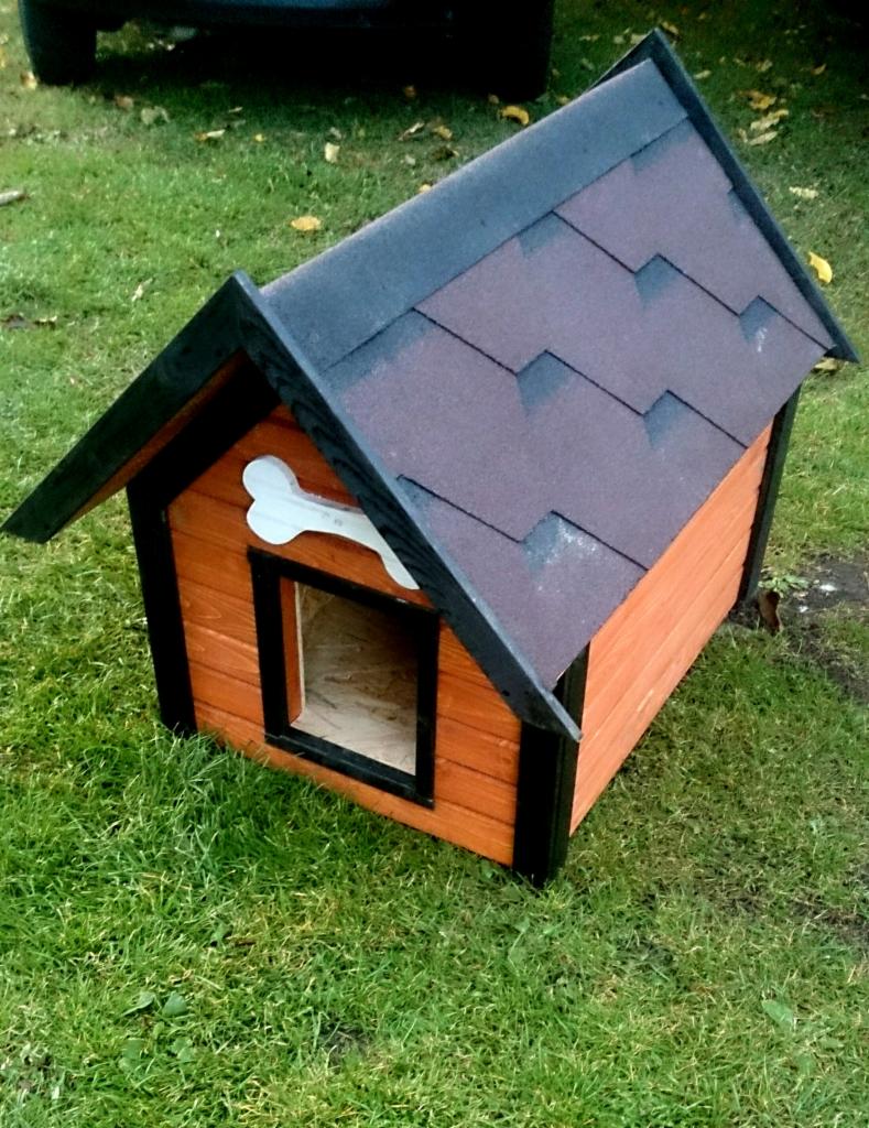 Drewniana buda dla psa z dachem krytym gontem bitumicznym, z dekoracyjną kością nad wejściem, wykonana w Kłobucku. Pomarańczowe ściany, czarne wykończenia.