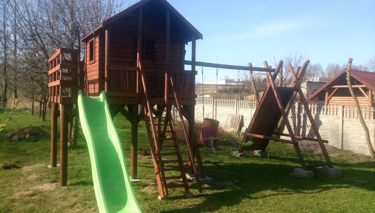 Drewniany plac zabaw z domkiem na palach, zjeżdżalnią i huśtawką na tle trawnika i ogrodzenia. Widoczna drabinka wejściowa i elementy konstrukcyjne. Słoneczny dzień.