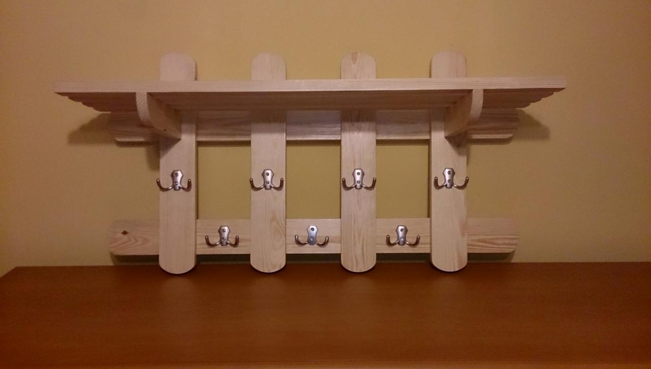 Drewniany wieszak z półką i metalowymi haczykami, wykonany w stylu rustykalnym. Jasne drewno, zaokrąglone krawędzie, widoczne słoje. Montowany na ścianie nad blatem.