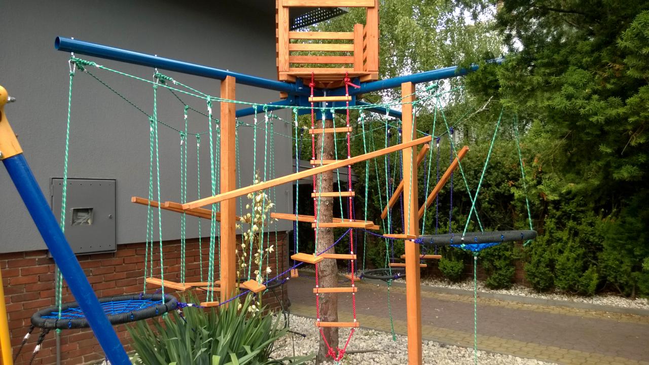 Drewniana konstrukcja placu zabaw z elementami linowymi i drabinką, wkomponowana w drzewo, z huśtawką na pierwszym planie i budynkiem w tle. Widok z poziomu gruntu.