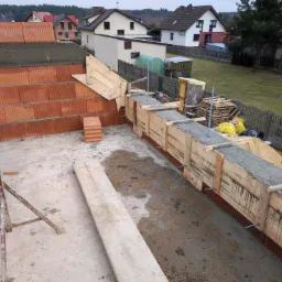 Wznoszenie ścian fundamentowych z bloczków ceramicznych, szalunek z desek zalewany betonem, widok z góry na plac budowy z cegłami i rusztowaniem w tle zabudowy jednorodzinnej.