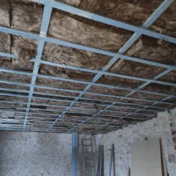 Sufit w trakcie izolacji wełną mineralną, widoczne metalowe profile konstrukcyjne, surowe ceglane ściany w tle, drabina oparta o ścianę.