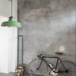 Szara ściana z efektem betonu w industrialnym wnętrzu, z rowerem opartym o ścianę, zieloną lampą wiszącą i stosem drewna przy ścianie. Minimalistyczny design z surowymi materiałami.