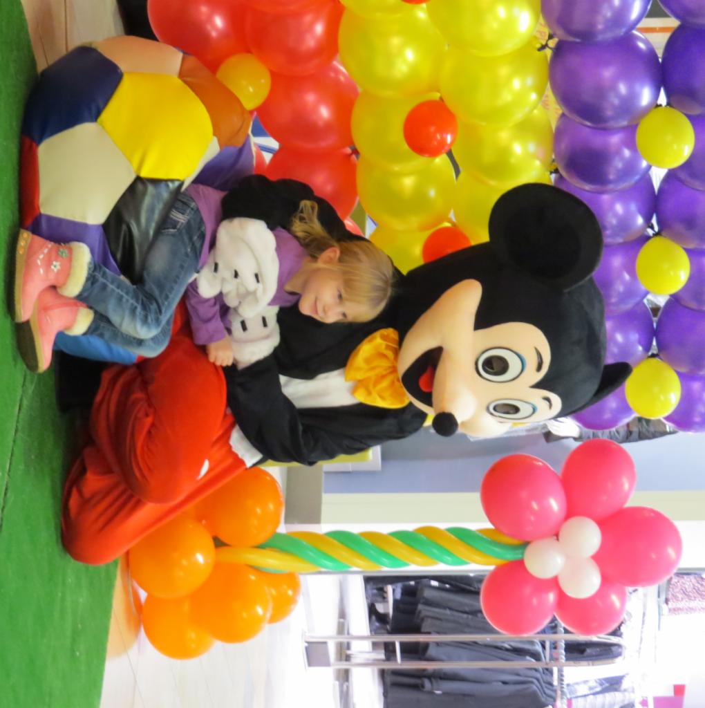 Dziewczynka w kostiumie Myszki Miki otoczona kolorowymi balonami: czerwonymi, żółtymi, fioletowymi i pomarańczowymi. Dodatkowo piłka i kwiat z balonów.
