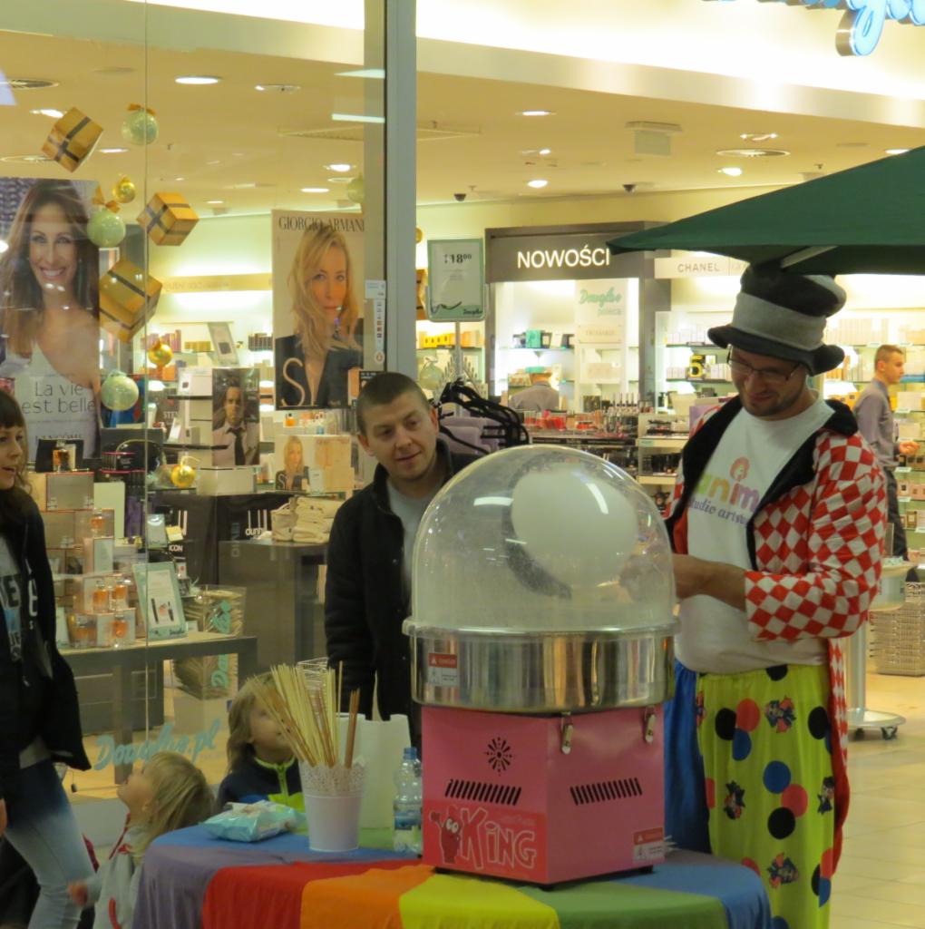 Animator w stroju klowna robi watę cukrową na tęczowym stole w centrum handlowym. W tle witryny sklepowe i osoby.
