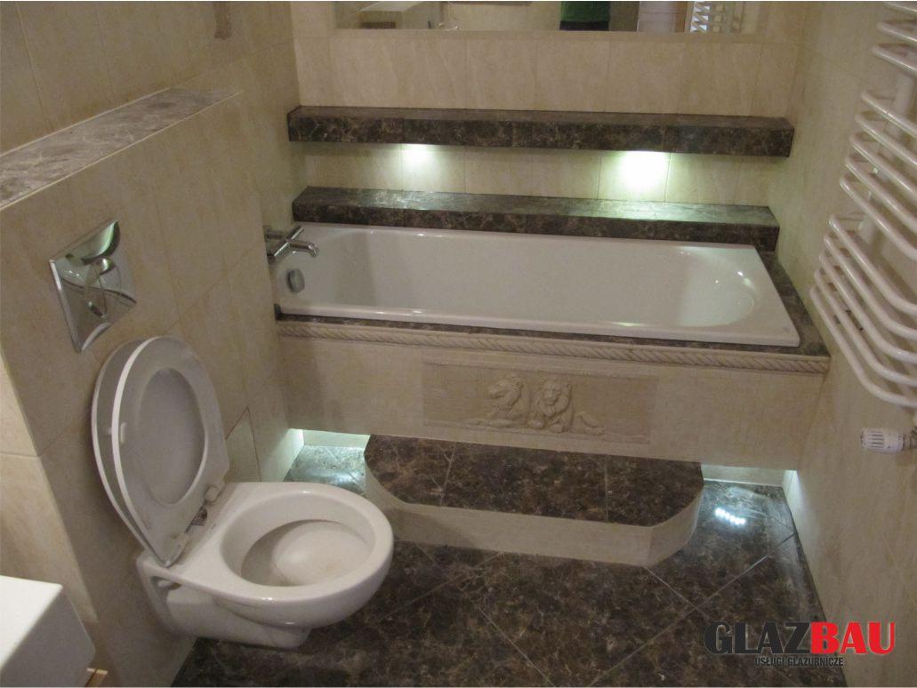 Beżowa łazienka z wanną obudowaną płytkami imitującymi marmur, podświetlona LED. Uwagę zwraca toaleta z otwartą klapą oraz dekoracyjne płaskorzeźby.