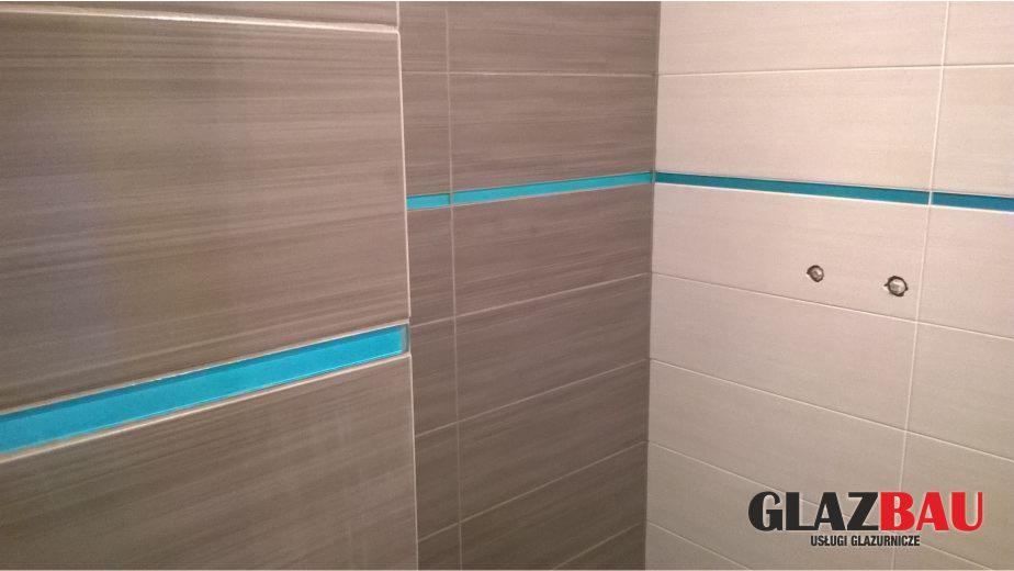 Wykończona ściana z płytek w odcieniach szarości, z poziomym paskiem dekoracyjnym w kolorze turkusowym. Widoczne otwory na armaturę i logo firmy glazurniczej.