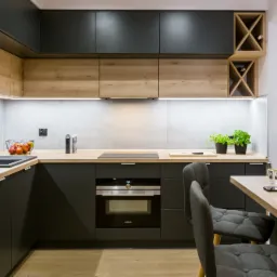 Nowoczesna kuchnia w ciemnych odcieniach szarości i drewna z elementami dekoracyjnymi: zioła w doniczkach, owoce w koszyku, w tle widoczna wnęka na wino.