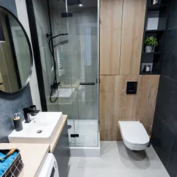 Nowoczesna łazienka z owalnym lustrem, kabiną prysznicową ze szkła i armaturą w kolorze czarnym, wykończona płytkami w odcieniach szarości i drewna.