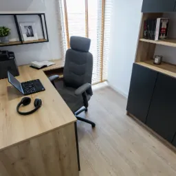 Nowoczesny gabinet w domu: biurko z laptopem, słuchawkami i kalendarzem, ergonomiczne krzesło, regał z książkami i dekoracjami.