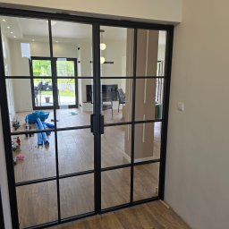 KONEKS Tomasz Kondera - Nowoczesne, dwuskrzydłowe drzwi wewnętrzne z czarnymi szprosami i przezroczystym szkłem, widok na salon w neutralnej kolorystyce, drewnopodobna podłoga.
