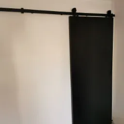 Czarna, przesuwna drzwi wewnętrzne na szynie, zamontowana w jasnym pomieszczeniu z szarą podłogą.