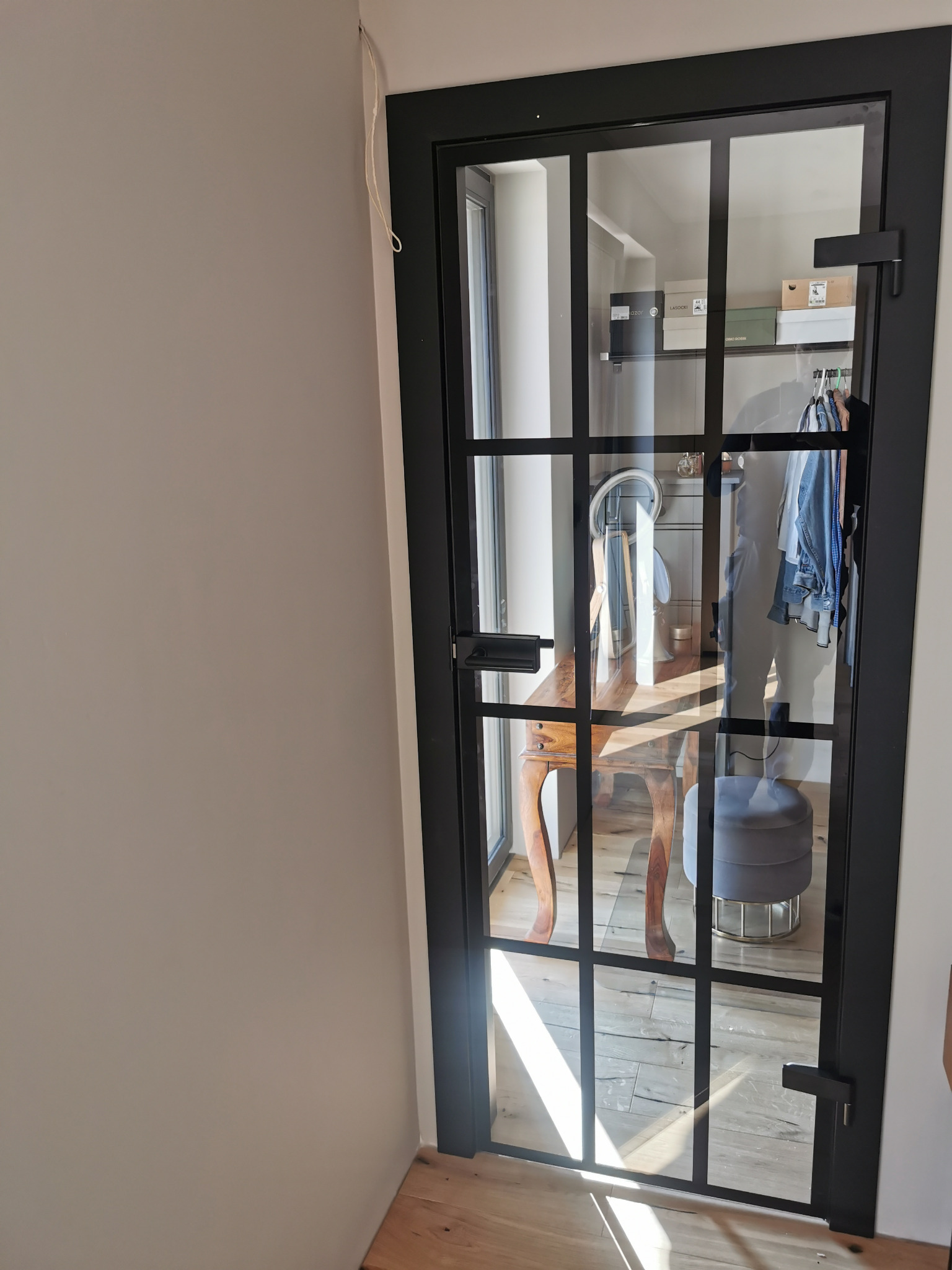 Szklane drzwi wewnętrzne z czarną ramą, dzielone na kwadraty, wpuszczające światło do garderoby z toaletką i pufą. Widok z boku, jasne wnętrze.