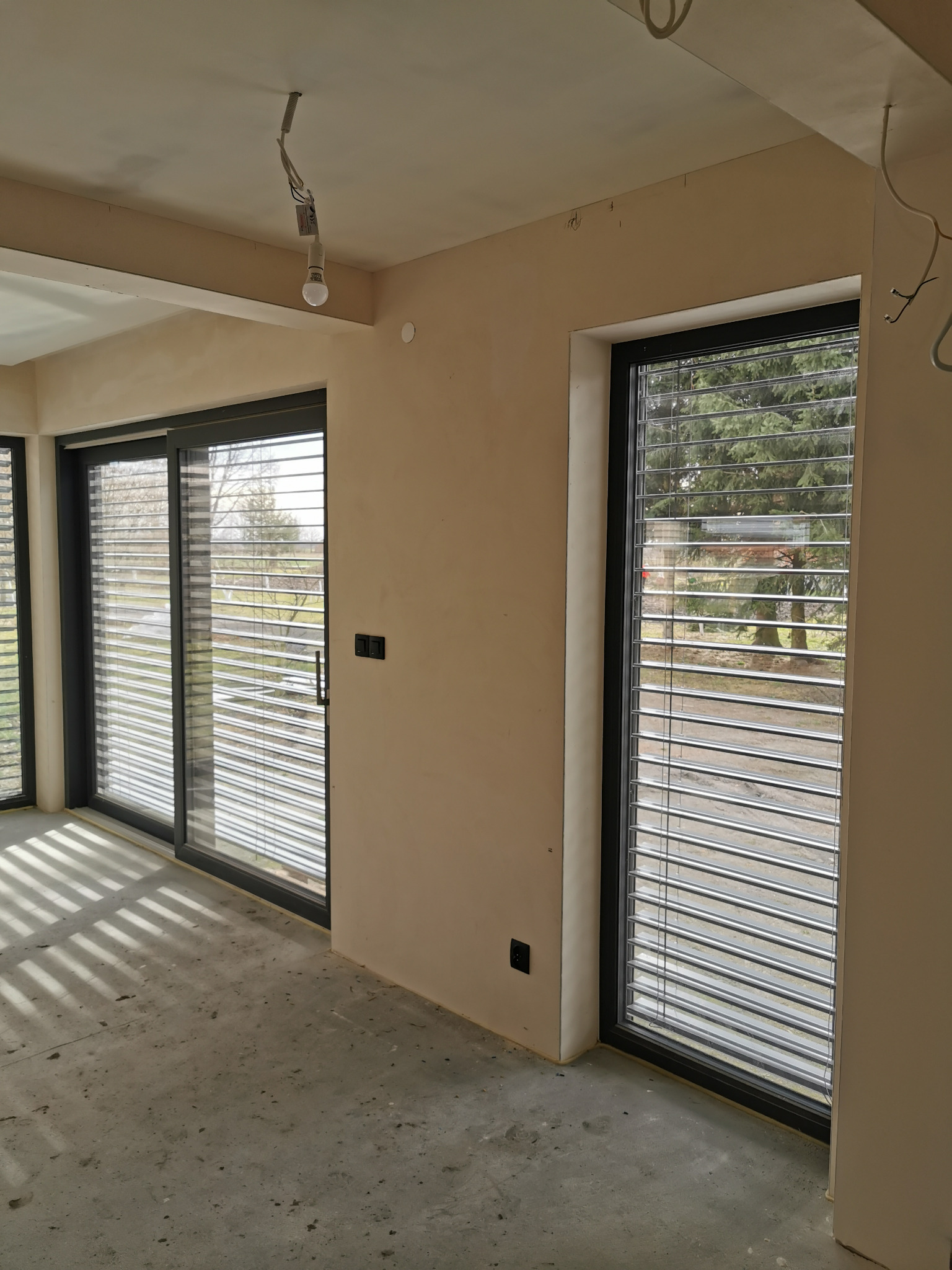 Wnętrze z dużymi oknami przesuwnymi z żaluzjami poziomymi w czarnych ramach. Surowy beton na podłodze, ściany w jasnym beżu, widoczne niedokończone instalacje.