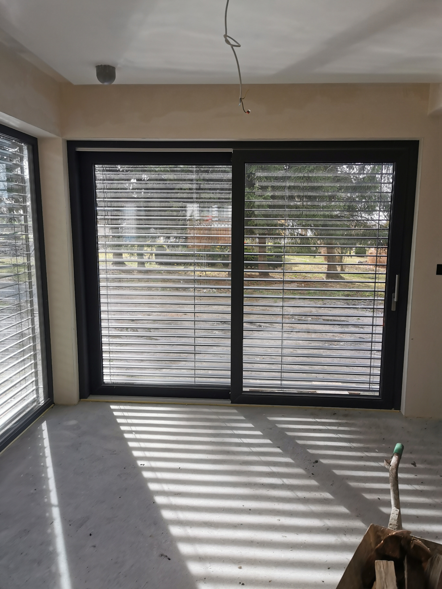 Duże, przesuwne okno z czarną ramą i żaluzjami wewnętrznymi, w surowym wnętrzu. Widoczne światło słoneczne wpadające przez żaluzje i taczka w rogu.