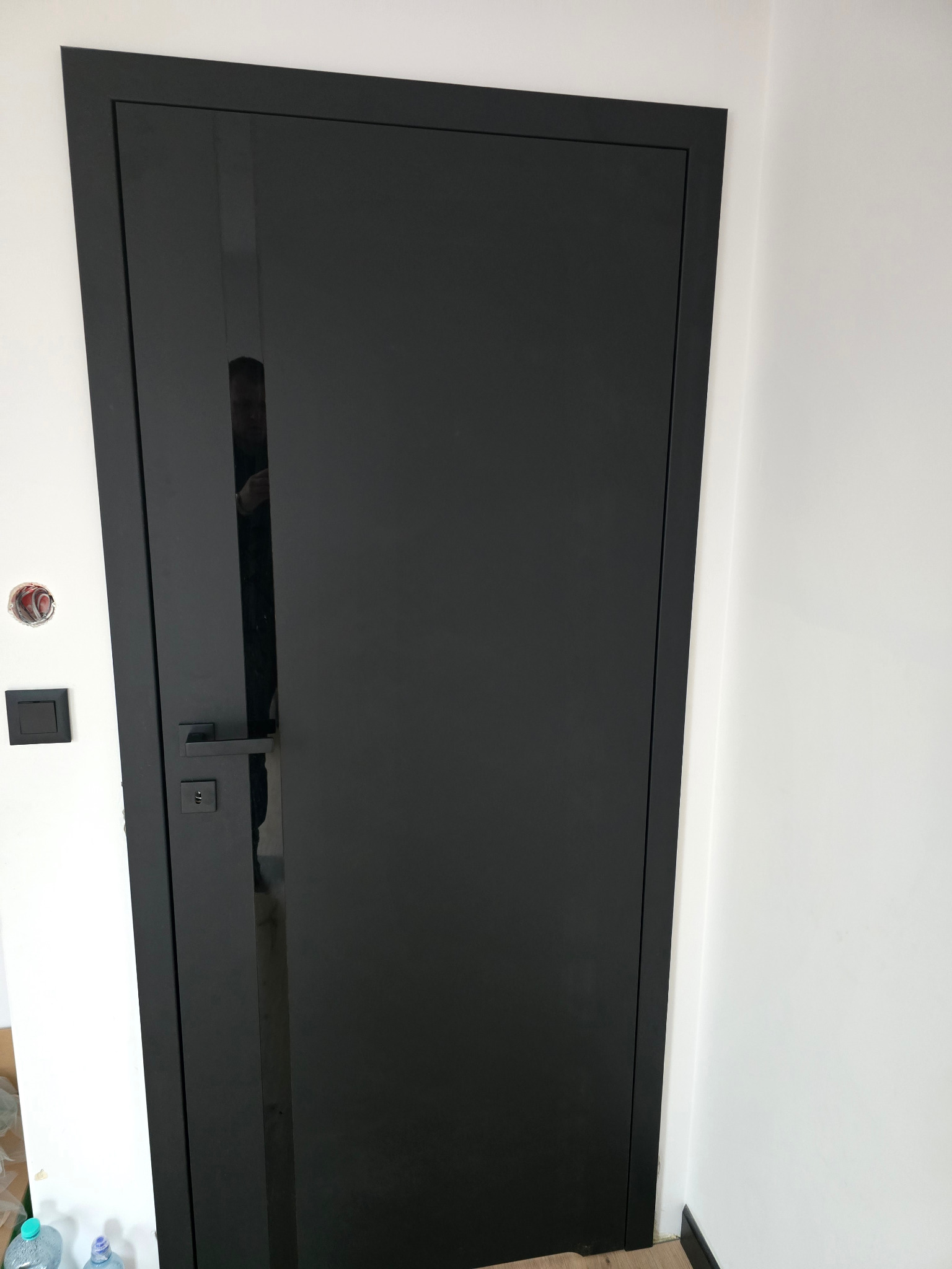 Czarne, matowe drzwi wewnętrzne z minimalistyczną klamką na tle białej ściany. Widoczny fragment instalacji elektrycznej i podłogi.