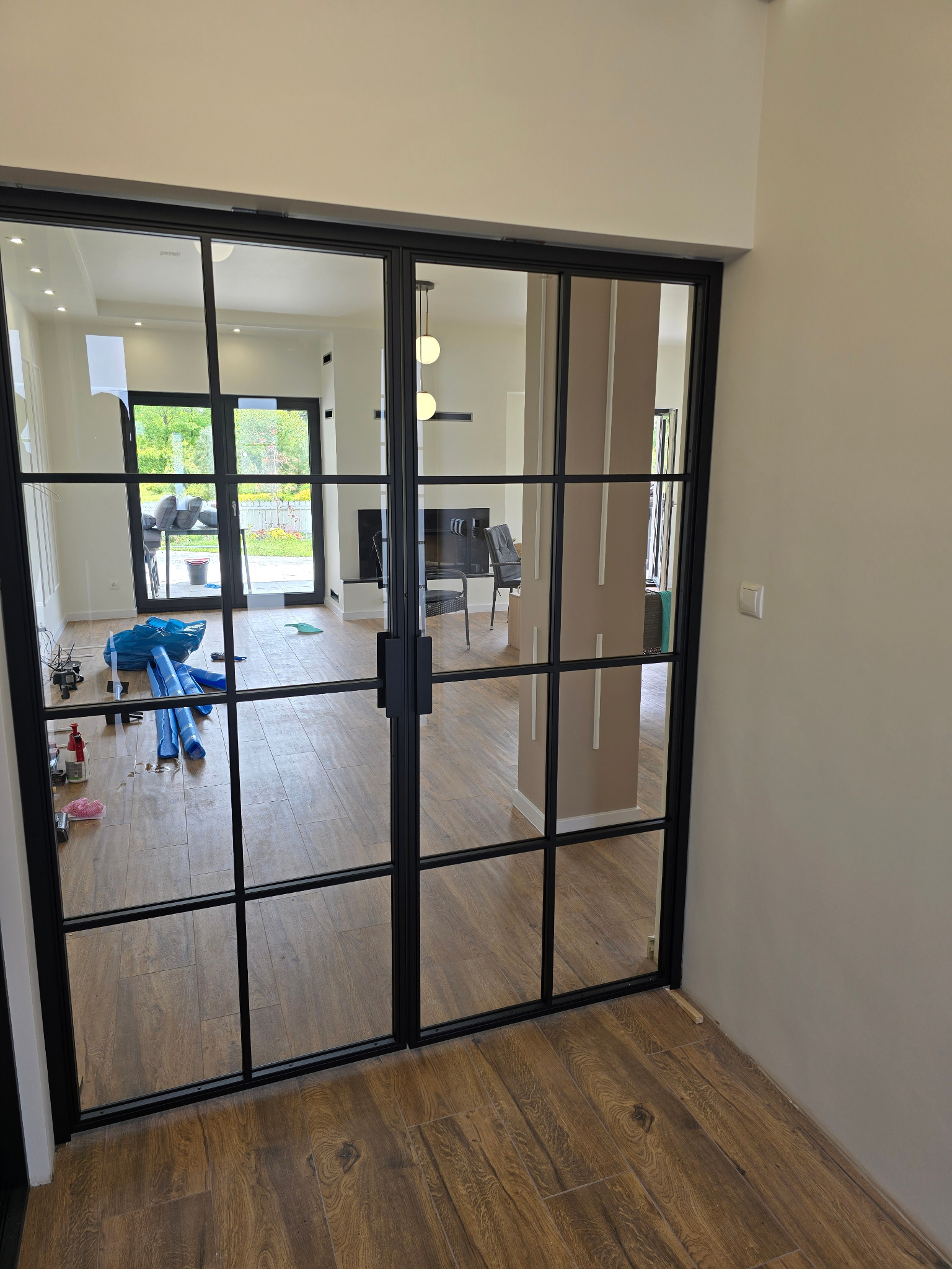 Nowoczesne, dwuskrzydłowe drzwi wewnętrzne z czarnymi szprosami i przezroczystym szkłem, widok na salon w neutralnej kolorystyce, drewnopodobna podłoga.