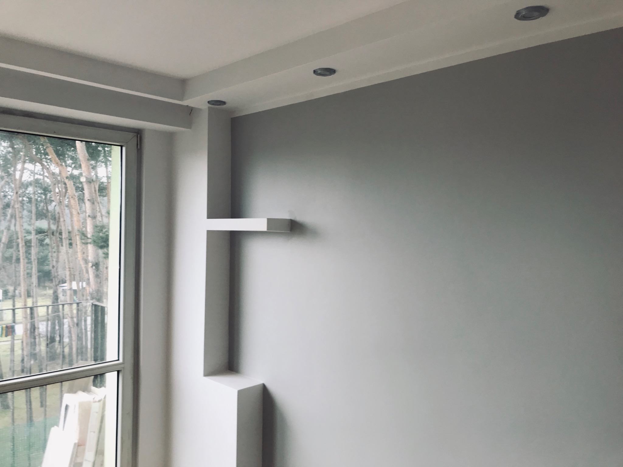 Szara ściana z zamontowanymi półkami w kształcie litery L, widok na zewnątrz przez okno balkonowe, widoczne drzewa i balustrada.