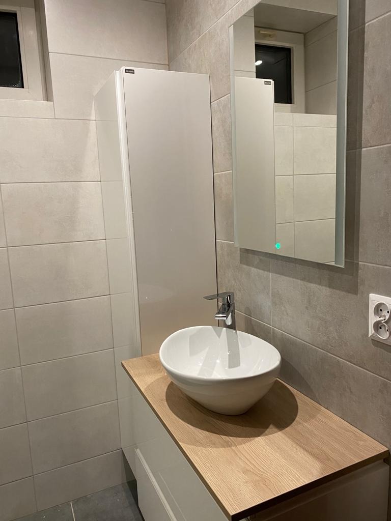 Nowoczesna łazienka z umywalką nablatową w kształcie misy, drewnianym blatem, białymi szafkami, dużym lustrem z podświetleniem LED i szarymi płytkami imitującymi kamień na ścianach.