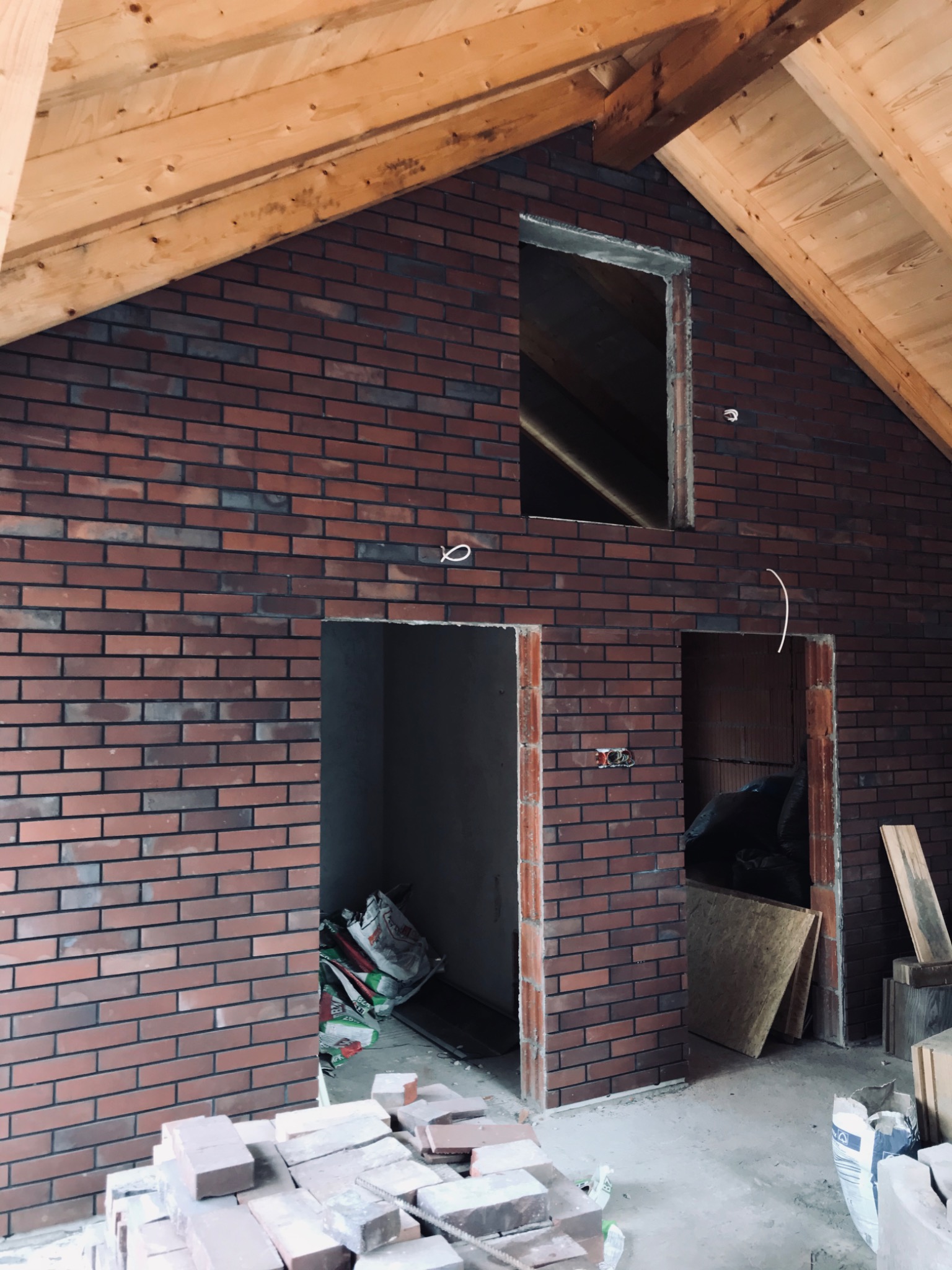 Murowana ściana z cegły klinkierowej w surowym stanie poddasza z widocznymi elementami konstrukcji drewnianej dachu, przygotowane otwory na okno i drzwi, widoczne przewody elektryczne i materiały...