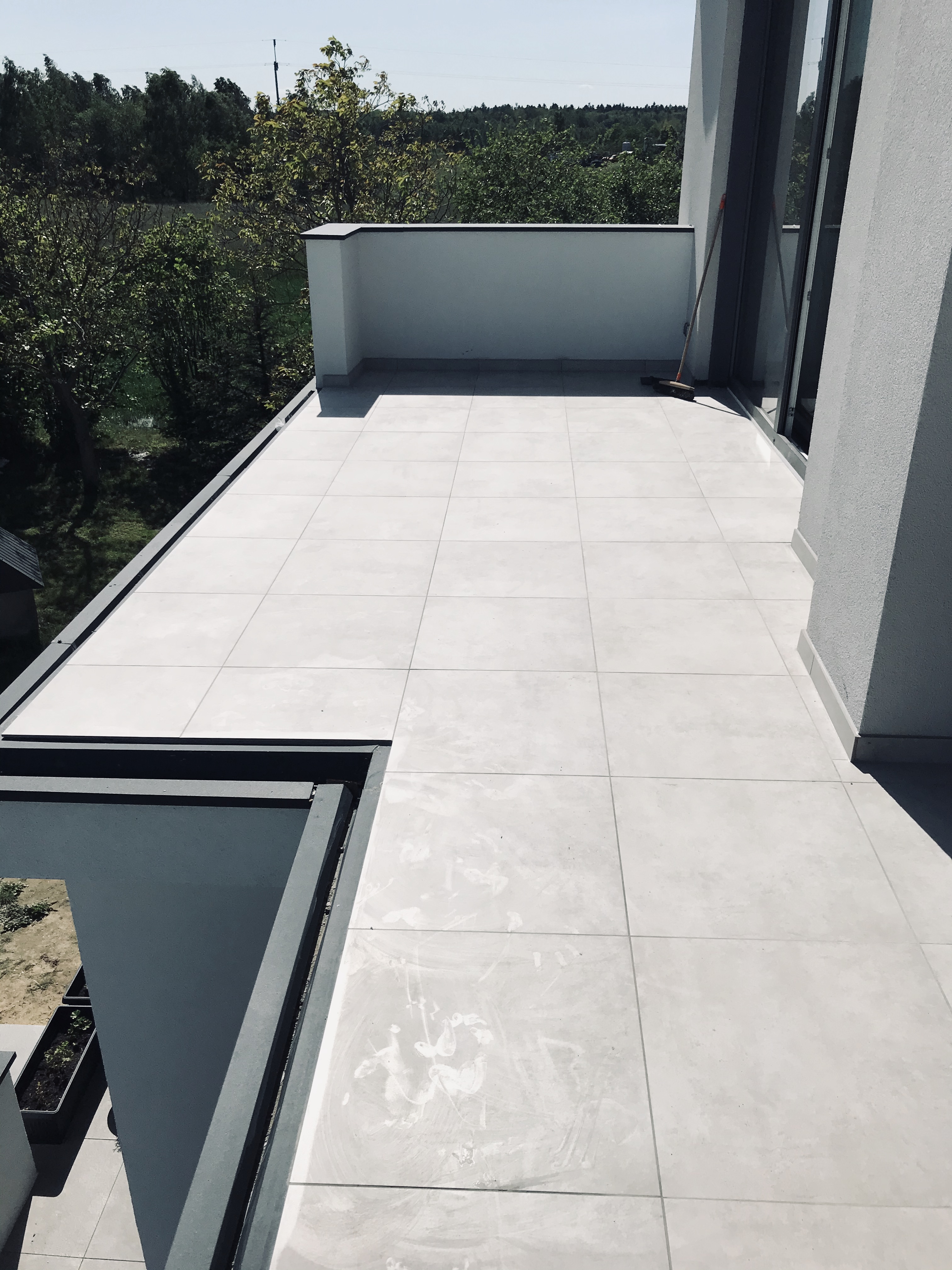 Wyłożony jasnymi płytkami taras z widokiem na zieleń, widoczna miotła oparta o ścianę, szare obramowanie tarasu.