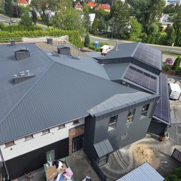 DACH-MAR Mariusz Adamczewski - Szary dach z blachy trapezowej, częściowo kryty, z panelami słonecznymi. Widok z góry na budynek, otoczenie miejskie z zielenią i fragmentem ulicy.
