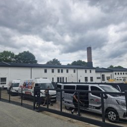 DACH-MAR Mariusz Adamczewski - Widok na biały budynek z ciemnym dachem, z kominem w tle i zaparkowanymi samochodami dostawczymi. Uwagę przyciąga brama z metalowych prętów na pierwszym planie.