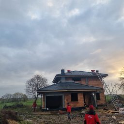 DACH-MAR Mariusz Adamczewski - Dom w trakcie budowy z czarnym dachem z dachówki, pracownicy w czerwonych ubraniach na dachu i wokół budynku. Pochmurne niebo w tle, krajobraz wiejski.