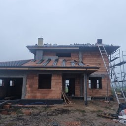 DACH-MAR Mariusz Adamczewski - Budowa domu jednorodzinnego w trakcie prac dekarskich: częściowo położone dachówki, ceglana konstrukcja, rusztowanie. Widok na budynek w pochmurny dzień.