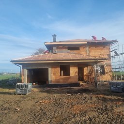 DACH-MAR Mariusz Adamczewski - Budowa domu jednorodzinnego w trakcie krycia dachu dachówką ceramiczną, widoczni dekarze pracujący na drewnianej konstrukcji, palety z dachówkami na ziemi.