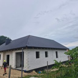 DACH-MAR Mariusz Adamczewski - Dom w trakcie budowy z ciemnym, nowym pokryciem. Widoczna osoba wchodząca na posesję. Tło stanowią drzewa i pochmurne niebo.