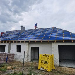 DACH-MAR Mariusz Adamczewski - Ekipa montuje konstrukcję dachu na nowym domu z bloczków Ytong. Widoczna niebieska membrana i drewniane łaty. Na pierwszym planie palety z dachówką.