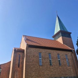 DACH-MAR Mariusz Adamczewski - Ceglany kościół z zieloną iglicą i krzyżem, widziany z dołu. Uwagę zwraca drabina oparta o ścianę, sugerująca prace konserwatorskie przy elewacji i pokryciu.