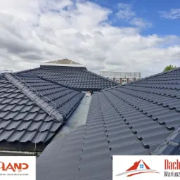 Czarny dach z dachówki ceramicznej w perspektywie z góry, na tle błękitnego nieba z chmurami. Widoczne logo firmy Dach-Mar. Dach po renowacji.