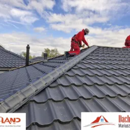 Dekarze w czerwonych kombinezonach montują ciemną dachówkę na skośnym dachu domu jednorodzinnego. Widoczne narzędzia i elementy wentylacyjne dachu na tle błękitnego nieba.