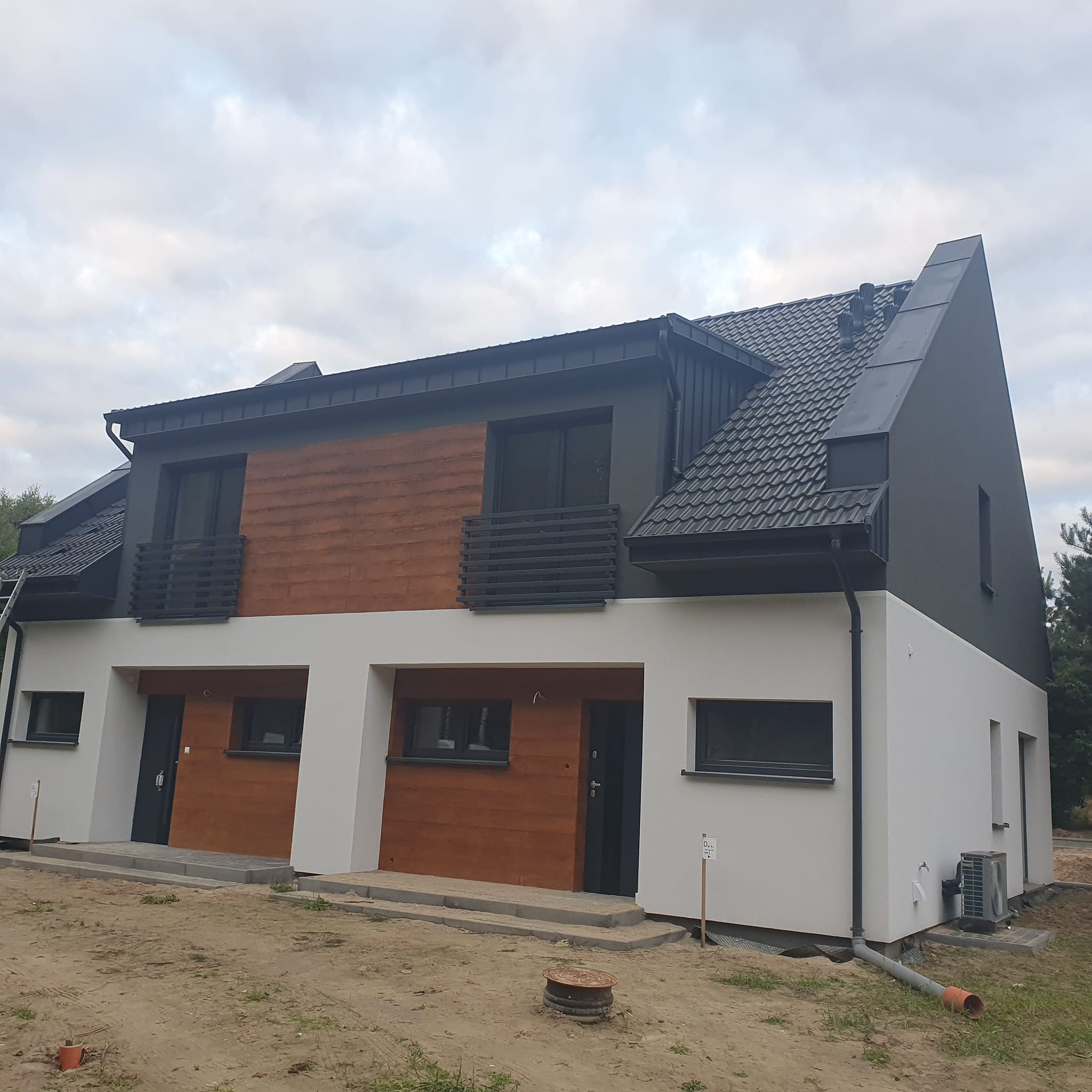 Nowoczesny bliźniak z ciemną dachówką karpiówką i orynnowaniem w kolorze grafitowym. Elewacja w dwóch odcieniach szarości z drewnianymi panelami. Widok z dołu, pochmurne niebo.
