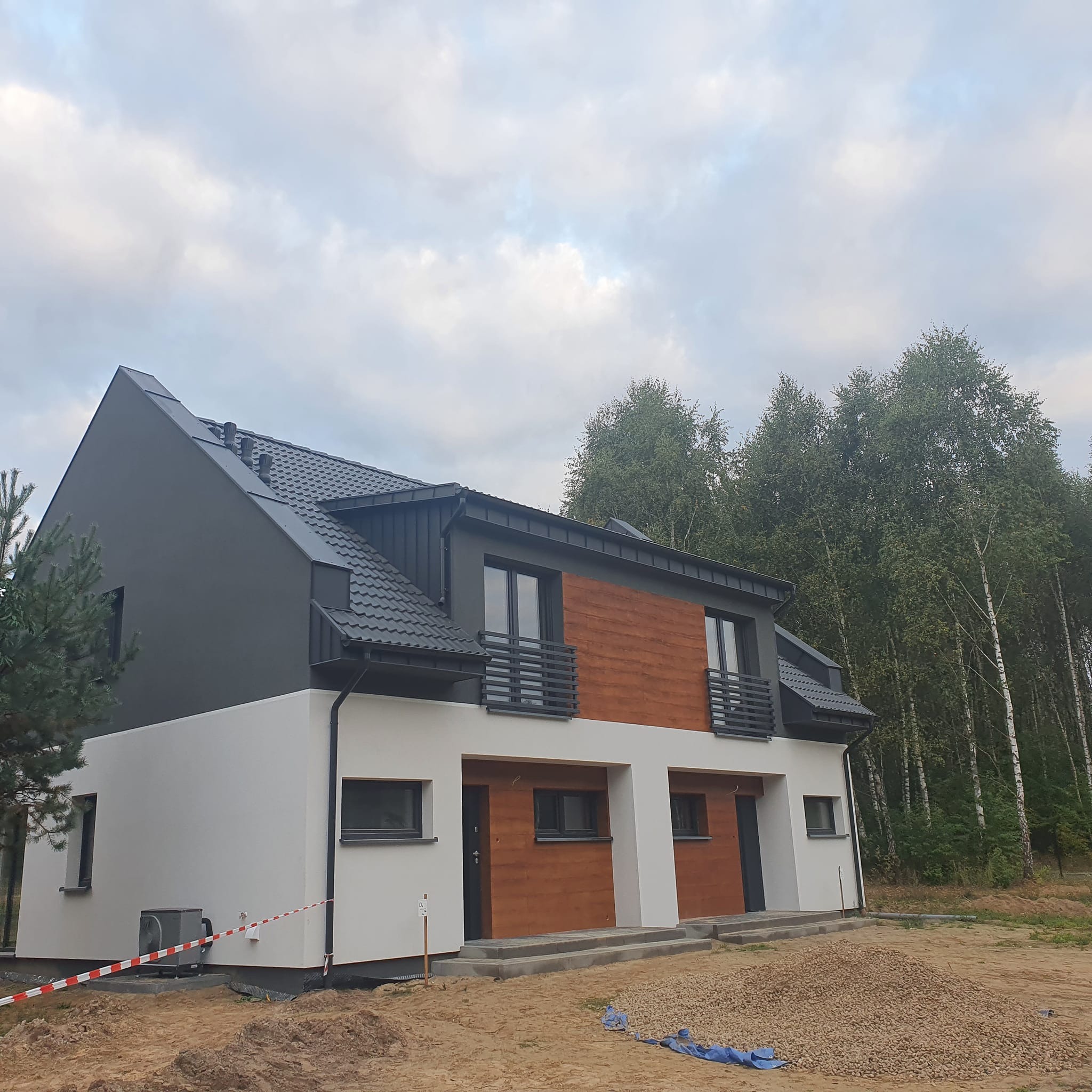 Nowoczesny dwukondygnacyjny dom bliźniak z ciemnoszarą dachówką i drewnianą elewacją. Widoczne rynny, okna i drzwi. W tle drzewa i pochmurne niebo.