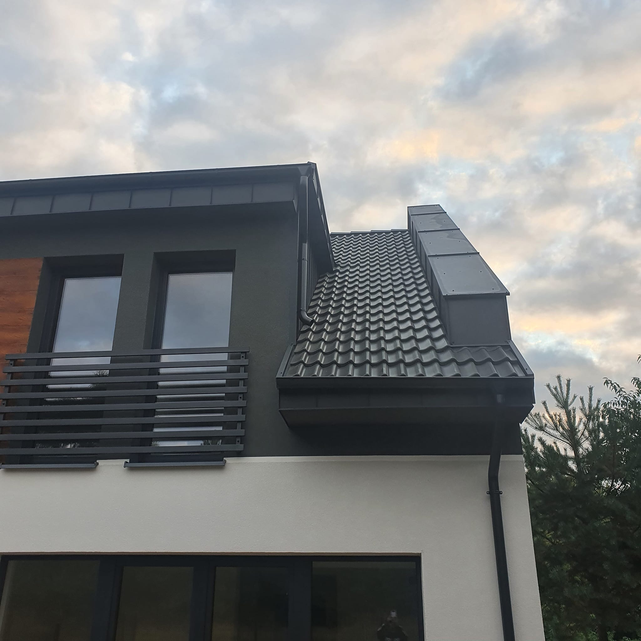 Nowoczesny dom z ciemną dachówką karpiówką i orynnowaniem, widok z dołu na tle pochmurnego nieba. Detal architektoniczny: komin obłożony blachą.