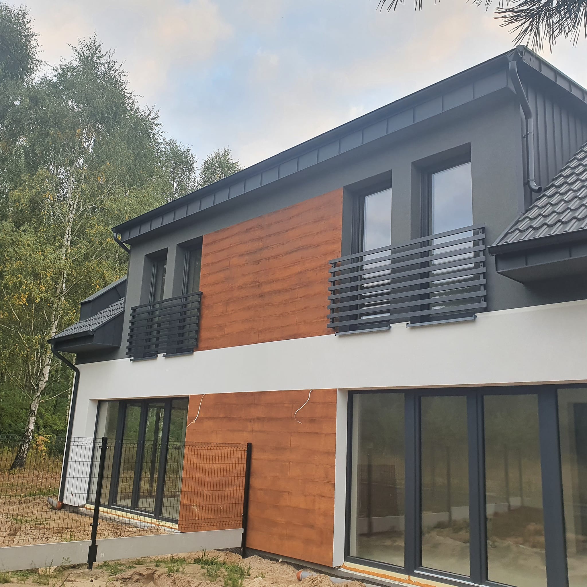 Nowoczesny dom dwupiętrowy z szarym dachem, elewacją w odcieniach szarości, brązu i bieli, z balkonami z metalowymi balustradami i dużymi oknami.